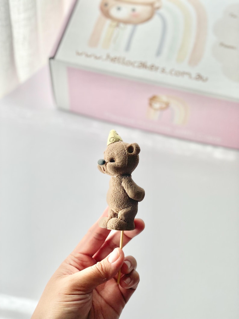 May include: A brown teddy bear cake topper with a textured surface, wearing a small yellow party hat on its head. The bear has a dark nose and is attached to a thin wooden stick for placement. The figure is shown in profile against a blurred box.
