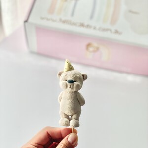 May include: A beige bear figurine topper with a textured surface, wearing a small yellow party hat with a dotted pattern. The bear has a dark gray nose and is attached to a thin wooden stick, positioned in front of a white box with a rainbow graphic.