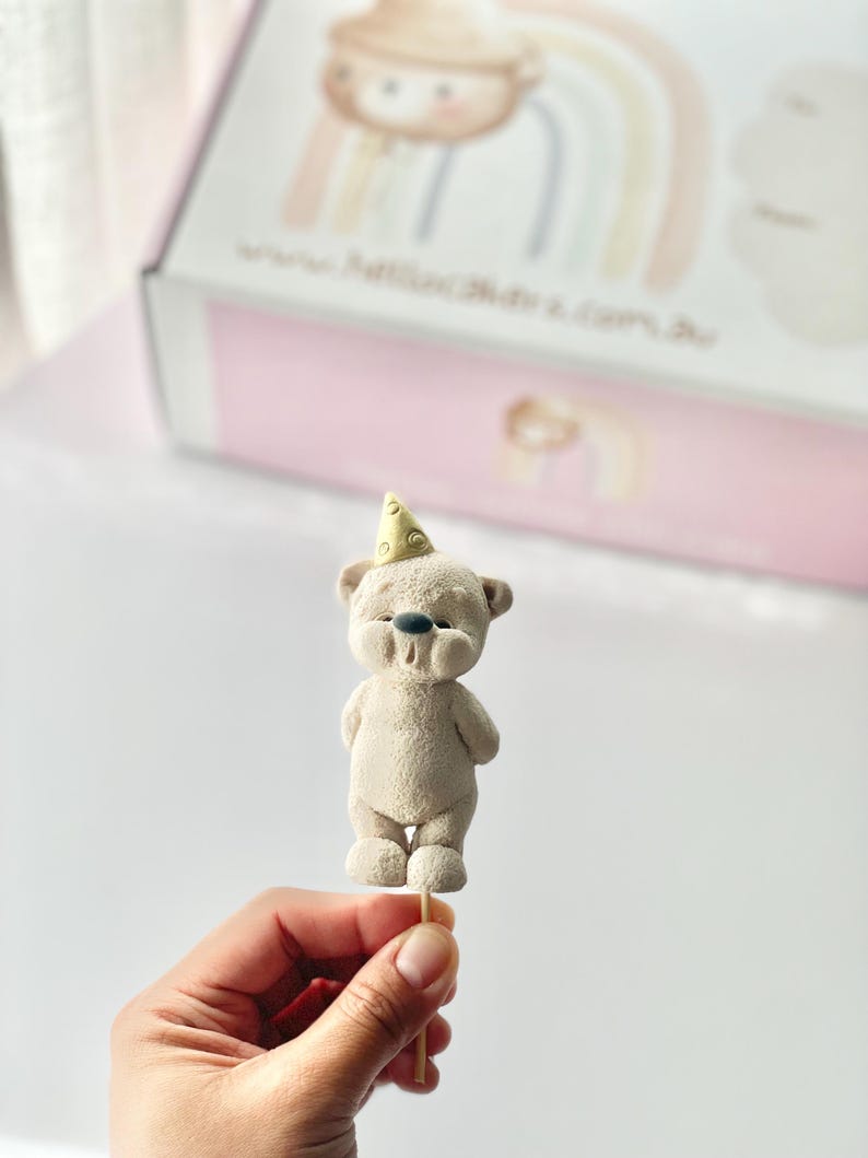 May include: A beige bear figurine topper with a textured surface, wearing a small yellow party hat with a dotted pattern. The bear has a dark gray nose and is attached to a thin wooden stick, positioned in front of a white box with a rainbow graphic.