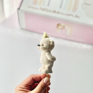 May include: A beige bear-shaped cake topper with a textured surface, wearing a small yellow party hat, mounted on a thin wooden stick. The bear is shown in profile, featuring a rounded body, small ears, and a dark nose, positioned upright.