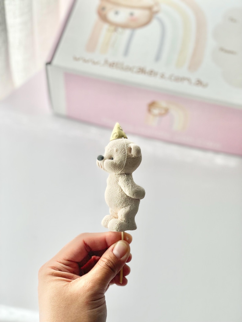 May include: A beige bear-shaped cake topper with a textured surface, wearing a small yellow party hat, mounted on a thin wooden stick. The bear is shown in profile, featuring a rounded body, small ears, and a dark nose, positioned upright.