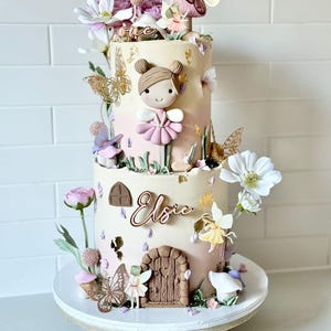 May include: A two-tiered cake decorated with a fairy theme. The cake is decorated with flowers, butterflies, and a small fairy figurine. The cake is also decorated with the name "Elsie" and the number "one".