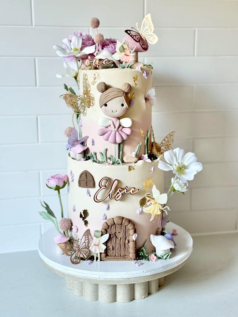 May include: A two-tiered cake decorated with a fairy theme. The cake is decorated with flowers, butterflies, and a small fairy figurine. The cake is also decorated with the name "Elsie" and the number "one".