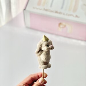 May include: A beige, textured, rabbit-shaped cake topper featuring a small, gold-colored party hat on its head. The rabbit is attached to a thin, wooden stick, and it has a rounded body, long ears, and a small pink nose detail.