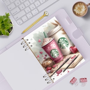 May include: A spiral-bound notebook featuring an illustration of a pink coffee cup and a white coffee cup, both with green logos, alongside a chocolate muffin, two pens, and scattered heart shapes. The notebook rests on a desk with a keyboard.