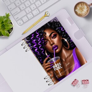 May include: A spiral-bound notebook featuring an illustration of a person with curly black hair with purple highlights, wearing a purple top and gold hoop earrings. The person holds a coffee cup with the text BOSS FUEL and a necklace reading Jesus.