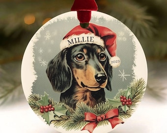 May include: A round Christmas ornament featuring a dachshund wearing a Santa hat with the name "Millie" and the year "2023". The ornament has a red ribbon for hanging and is decorated with holly and a red bow.