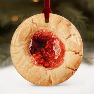 May include: A round ceramic ornament featuring a realistic print of a thumbprint cookie with a golden-brown, textured dough edge and a vibrant, glossy red jam center. The ornament hangs from a dark red velvet ribbon loop at the top.