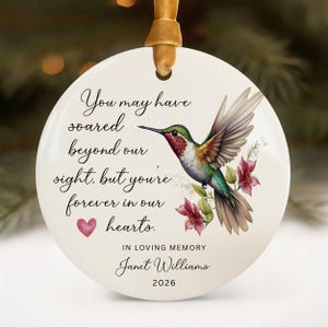 May include: A white ceramic memorial ornament with a hummingbird illustration and the text "You may have soared beyond our sight, but you're forever in our hearts." Includes the name Janet Williams and the year 2026.