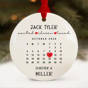 May include: A white, round Christmas ornament with a red ribbon. The ornament features the names "JACK TYLER" and "MILLER", along with the words "wanted, chosen, loved". A calendar highlights October 15, 2026.