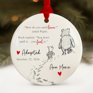 May include: A round white ceramic ornament with a red ribbon, featuring a black ink illustration of Winnie the Pooh and Piglet. Text reads: How do you spell love? asked Piglet. Pooh replied, You don't spell it... you feel it. Adopted October 22, 2026, Ava Marie.
