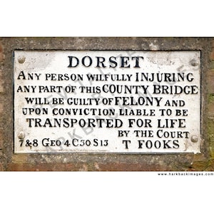 May include: A weathered rectangular sign with the word "DORSET" at the top. The sign has text warning against damaging the county bridge, with the phrase "TRANSPORTED FOR LIFE." The sign is mounted on a brick wall.