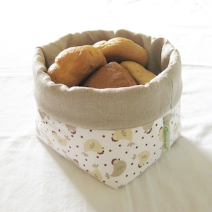Op de afbeelding: Een beige linnen broodmand met een witte en bruine kip print. De mand is gevuld met verschillende broodjes.