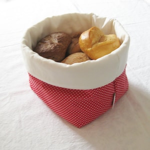 May include: A fabric bread basket with a white interior and a red and white polka dot exterior. The basket contains several baked rolls and bread. The basket is designed to hold bread and keep it warm.