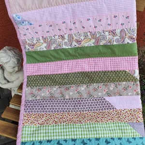 Könnte beinhalten: Ein Patchwork-Quilt mit verschiedenen Stoffen in Rosa-, Grün- und Blautönen. Der Quilt hat Blumen-, Paisley- und Vichy-Muster sowie einen gepunkteten rosa Rand. Der Quilt ist etwa 76 cm lang und 51 cm breit.