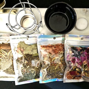 May include: An assortment of items for a spiritual practice. Includes four clear, sealed bags filled with herbs and botanicals, a silver metal candle holder, a black bowl, and a white candle. The items are arranged on a white and black surface.