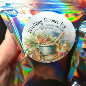 Puede incluir: Una bolsa holográfica con una etiqueta redonda que dice "Holiday Simmer Pot" con una imagen de una olla, ramitas de pino y regalos. La bolsa está atada con una cinta roja y verde. La bolsa contiene una botella de poción brillante.