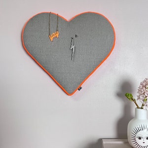 Könnte beinhalten: Ein graues herzförmiges Pinnwand mit orangefarbenem Rand. Die Pinnwand hat eine Halskette mit einem "you"-Anhänger und einem Blitzanhänger. Eine weiße Vase mit einem Sonnen-Gesicht-Design hält rosa Blumen. Zwei orangefarbene Textmarker liegen auf einer weißen Oberfläche.