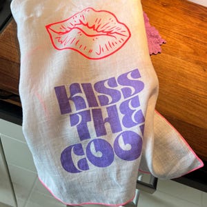 May include: A white linen tea towel with a pink outline. The towel has a purple graphic that says "Kiss the Cool" and a red outline of a pair of lips.