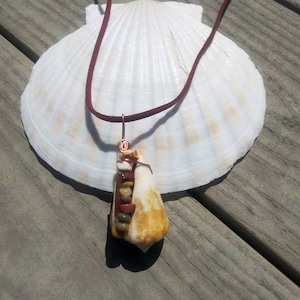 May include: A handmade necklace featuring a pendant made from a seashell and small, colorful stones. The pendant is attached to a dark red cord. The necklace rests on a large, white seashell, set against a weathered wooden surface.