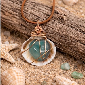 May include: A handcrafted necklace featuring a teal-colored stone encased in copper wire, set within a seashell. The pendant hangs from a brown leather cord. The necklace is displayed on a sandy surface with seashells and driftwood.