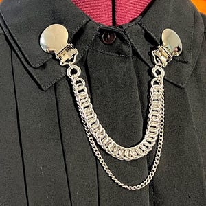 Könnte beinhalten: Silberfarbene Kragenclips mit Kettenbesatz, befestigt an einem schwarzen Hemd mit Kragen. Die Clips haben eine abgerundete Form und werden mit einem Verschluss am Hemd befestigt. Die Kette besteht aus kleinen, ineinandergreifenden Ringen.