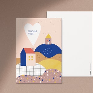 Puede incluir: Una postal rosa y blanca con forma de coraz&oacute;n y el texto "Sending Hugs". La postal presenta una ilustraci&oacute;n colorida de dos casas en colinas con un esquema de color azul, amarillo y marr&oacute;n.