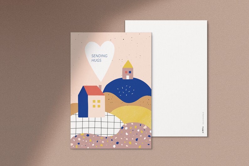 Puede incluir: Una postal rosa y blanca con forma de coraz&oacute;n y el texto "Sending Hugs". La postal presenta una ilustraci&oacute;n colorida de dos casas en colinas con un esquema de color azul, amarillo y marr&oacute;n.