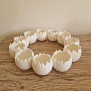 May include: Eleven white, eggshell-shaped containers with a jagged edge, arranged in a circle. The objects sit on a wooden surface. The design suggests a craft or decorative theme. The neutral colour palette and unique shapes create a minimalist aesthetic.