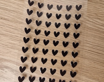 May include: A sheet of small, black heart-shaped stickers arranged in neat rows. The stickers are on a clear, rectangular backing, set against a light brown wooden surface. The hearts have a glossy finish.