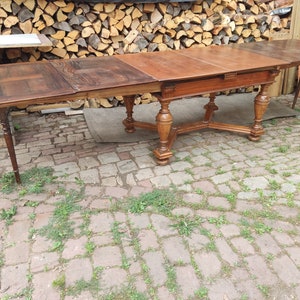 Könnte beinhalten: Ein großer, ausziehbarer Esstisch aus Holz mit aufwendigen Schnitzereien an den Beinen. Der Tisch ist aus dunklem Holz und hat eine rechteckige Form. Der Tisch steht auf einer Terrasse aus Ziegeln.