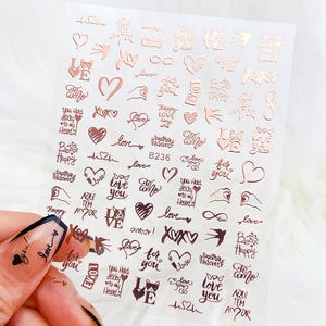 May include: A sheet of rose gold metallic nail art stickers featuring various romantic designs including hearts, swallows, infinity symbols, and text phrases like love, you hold the key to my heart, and te amo, with the model number B236 printed.