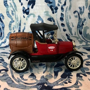 Op de afbeelding: Een rode vintage speelgoedauto met een zwart dak en zilveren wielen. De auto heeft een bruin vat aan de achterkant met de tekst "True Value". De auto heeft ook het nummer "8" aan de zijkant.