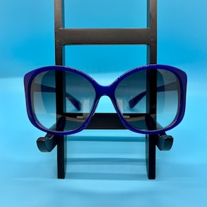 May include: Blue sunglasses with a dark gradient lens. The sunglasses have a thick, rounded frame and are displayed on a black stand against a blue background. The sunglasses are a vibrant shade of blue.