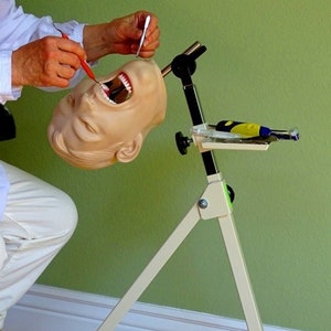 May include: A white dental training mannequin head mounted on a white tripod stand with a tray for dental tools. The mannequin head is open-mouthed and has realistic teeth. The stand has a black arm with a green knob. The text "Copyright Buyamag Inc Dental Education Supply 760-804-1434" is visible on the stand.