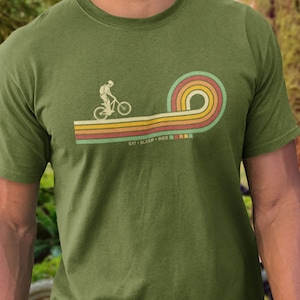 May include: A green t-shirt with a retro-style graphic of a person riding a bicycle. The graphic is in shades of orange, yellow, and blue. The text "Eat Sleep Ride" is printed below the graphic.