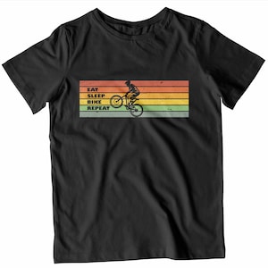 May include: Black t-shirt with a cycling graphic. The design shows a cyclist doing a wheelie, with the words "EAT SLEEP BIKE REPEAT" above. The background has horizontal stripes in orange, yellow, green, and brown.