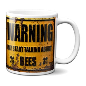 May include: White ceramic mug featuring a yellow and brown graphic with the text "WARNING MAY START TALKING ABOUT BEES". The design also includes two bee illustrations in black.
