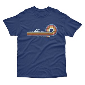 May include: Navy blue t-shirt with a retro-style graphic of a swimmer in a wave of colorful stripes. The text "EAT SLEEP SWIM" is printed below the graphic.