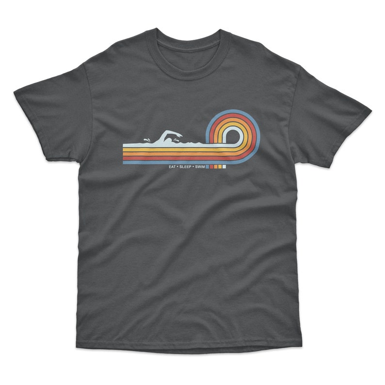 May include: A dark gray t-shirt with a retro-style graphic of a swimmer in a wave. The graphic is in shades of orange, yellow, blue, and red. The text "EAT SLEEP SWIM" is printed below the graphic.