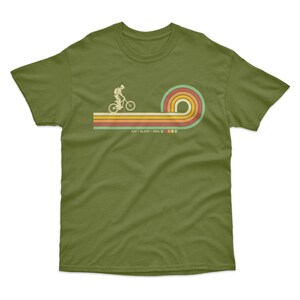 May include: A green t-shirt with a retro-style graphic of a person riding a mountain bike on a rainbow-colored path. The text "EAT + SLEEP + RIDE" is printed below the graphic.