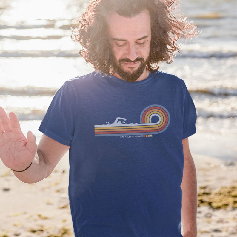 May include: A blue t-shirt with a retro-style graphic of a swimmer in a wave. The graphic is in shades of orange, yellow, and blue. The text "Eat - Sleep - Swim" is printed below the graphic.