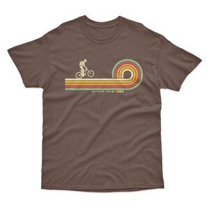 May include: A brown t-shirt with a retro-style graphic of a person riding a bicycle over a rainbow. The text "EAT + SLEEP + RIDE" is printed below the graphic.