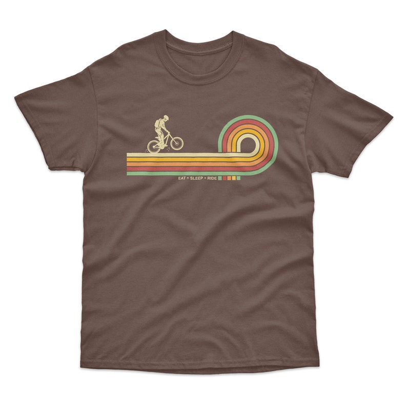 May include: A brown t-shirt with a retro-style graphic of a person riding a bicycle over a rainbow. The text "EAT + SLEEP + RIDE" is printed below the graphic.