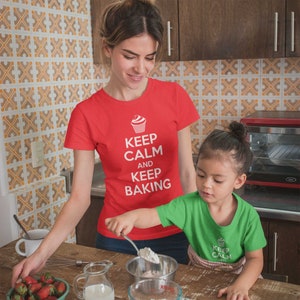 May include: A woman and a child wearing matching red and green t-shirts with a cupcake graphic and the text "Keep Calm and Keep Baking". They are in a kitchen and are baking together.