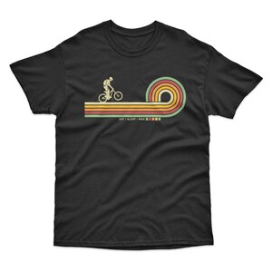 May include: Black t-shirt with a retro-style graphic of a person riding a bicycle on a rainbow-colored path. The text "EAT - SLEEP - RIDE" is below the graphic.