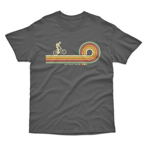 May include: A dark gray t-shirt with a retro-style graphic of a person riding a mountain bike over a rainbow. The text "EAT + SLEEP + RIDE" is printed below the graphic.