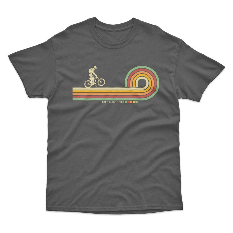 May include: A dark gray t-shirt with a retro-style graphic of a person riding a mountain bike over a rainbow. The text "EAT + SLEEP + RIDE" is printed below the graphic.