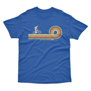 May include: A royal blue t-shirt with a retro-style graphic of a person riding a bicycle on a rainbow-colored stripe. The text "EAT + SLEEP + RIDE" is printed below the graphic.