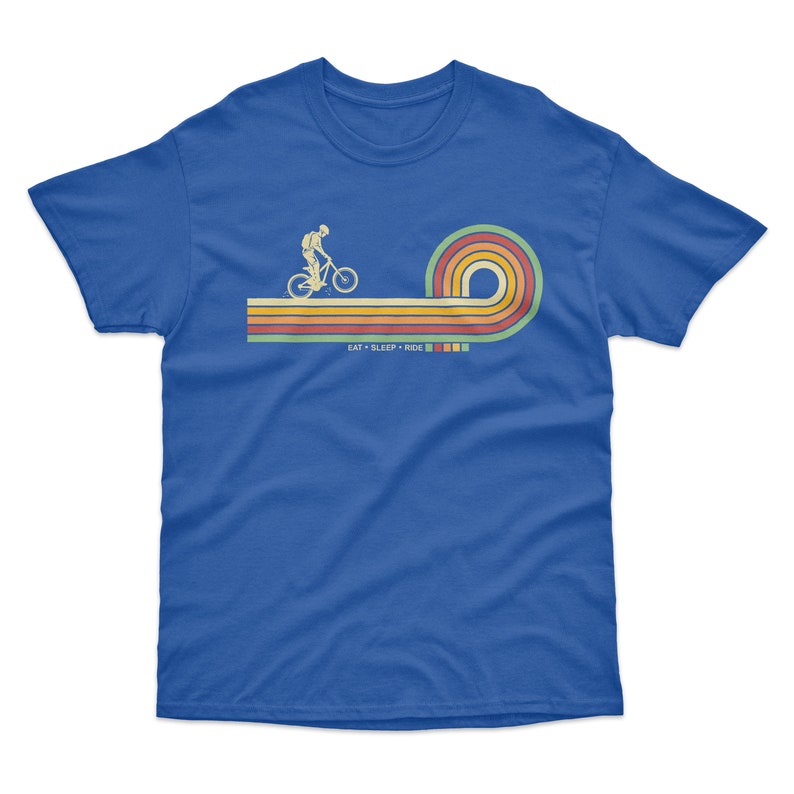 May include: A royal blue t-shirt with a retro-style graphic of a person riding a bicycle on a rainbow-colored stripe. The text "EAT + SLEEP + RIDE" is printed below the graphic.